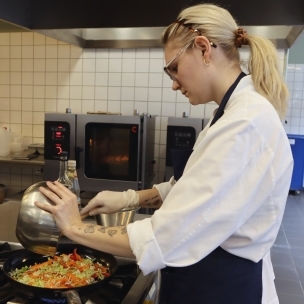Yngre kvinde i storkøkken hælder grøntsager ned i en pande 
